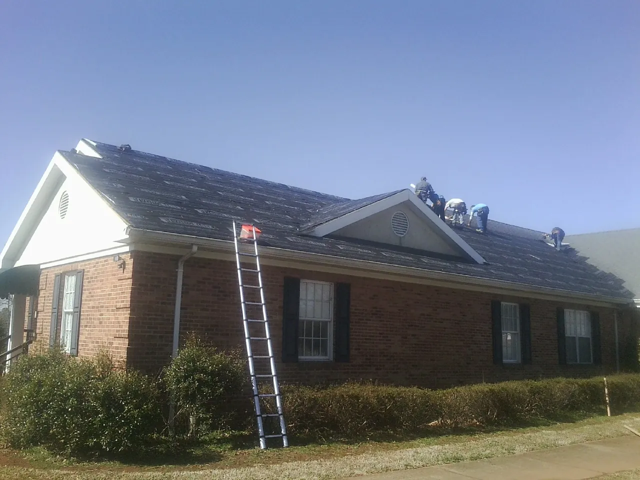 Crew tearing shingles off a brick ranch roof with a ladder set — Metal Roof Repair work by our crew in Elkmont
