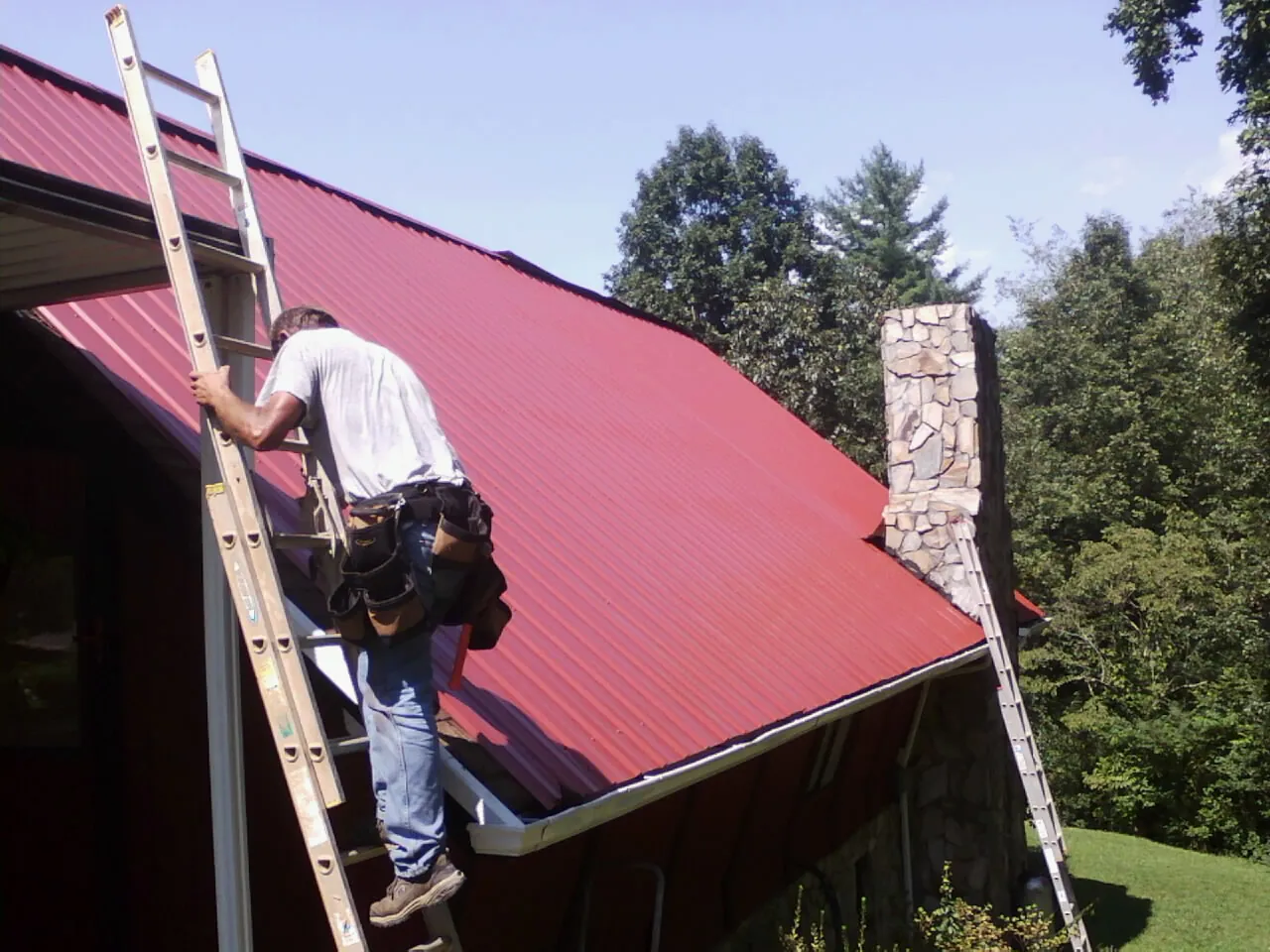 Roofer on a ladder beside a red metal roof and stone chimney — Soffit & Fascia Repair by Roofing Brothers in Elkmont